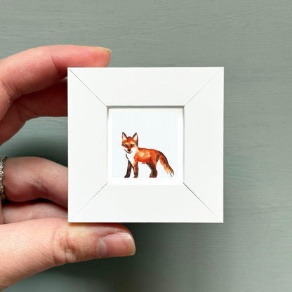 A hand is holding a small white square frame containing a picture of a fox.