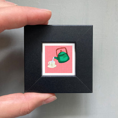 A hand is holding a small black square frame containing an illustration of a green teapot.
