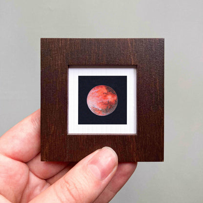 A hand is holding a small wooden frame containing a framed image of a red planet, likely Mars, against a dark background.