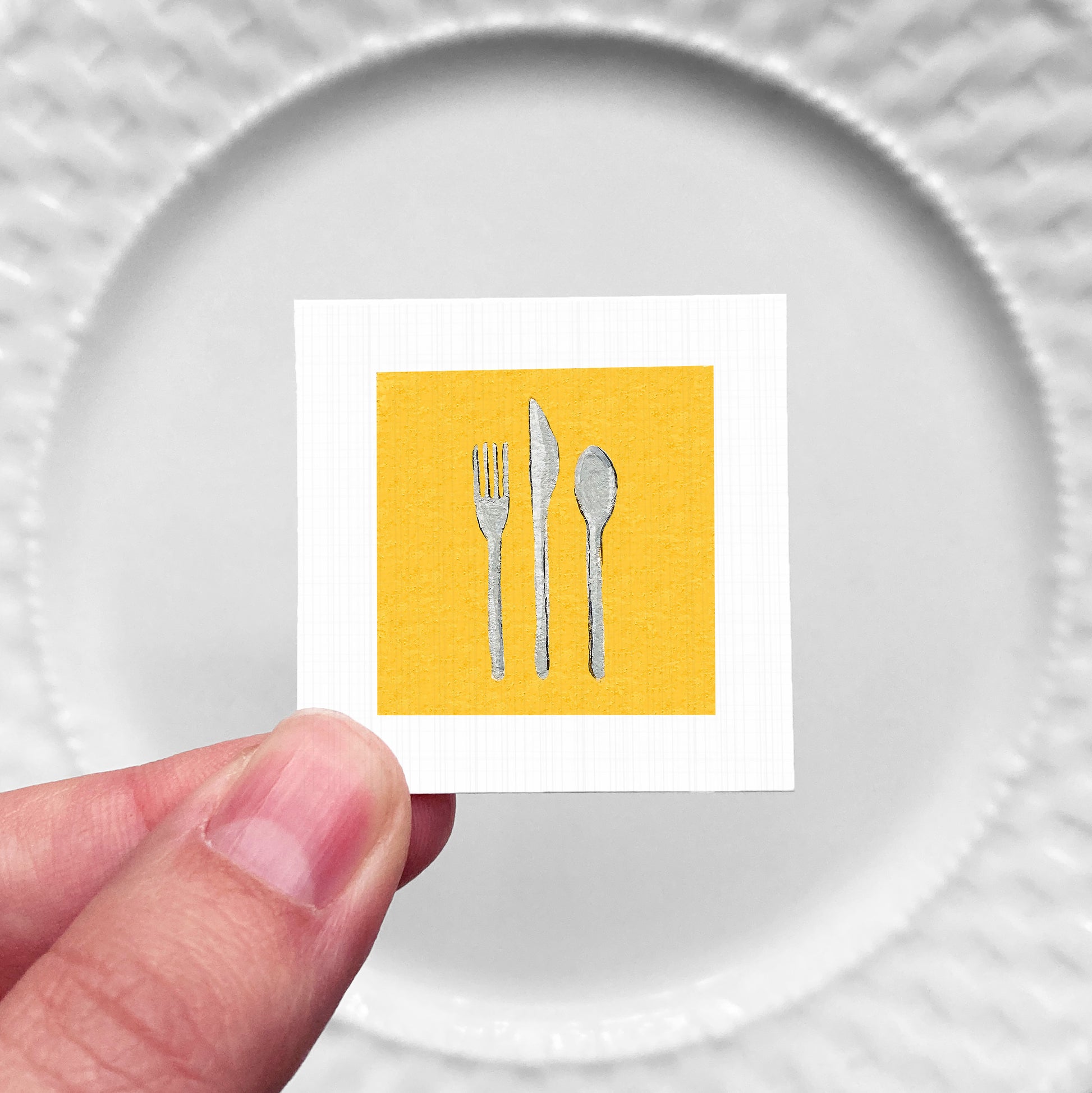 A hand is holding a small square with a yellow background, featuring a picture of a fork, knife, and spoon.