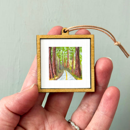 A hand holding a small wooden frame containing a painting of a forest road.