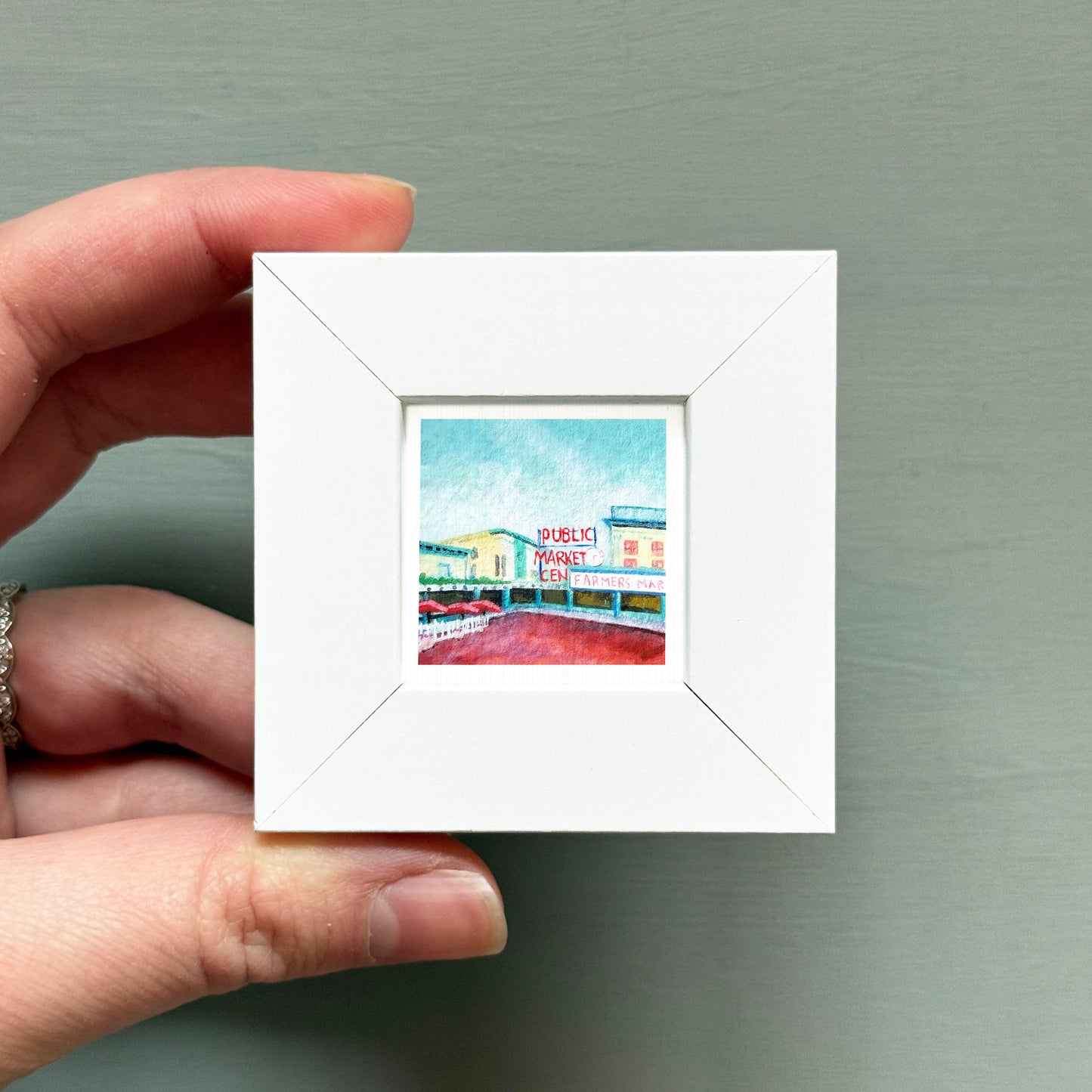 A hand is holding a small white square frame containing a painting of a building with a sign that reads "BAR & LOBBY".