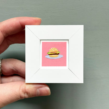 A hand is holding a small white picture frame containing an illustration of a hamburger.