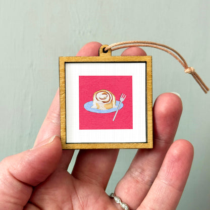 A person's hand holding a small wooden frame containing a framed image of a cinnamon roll on a plate.