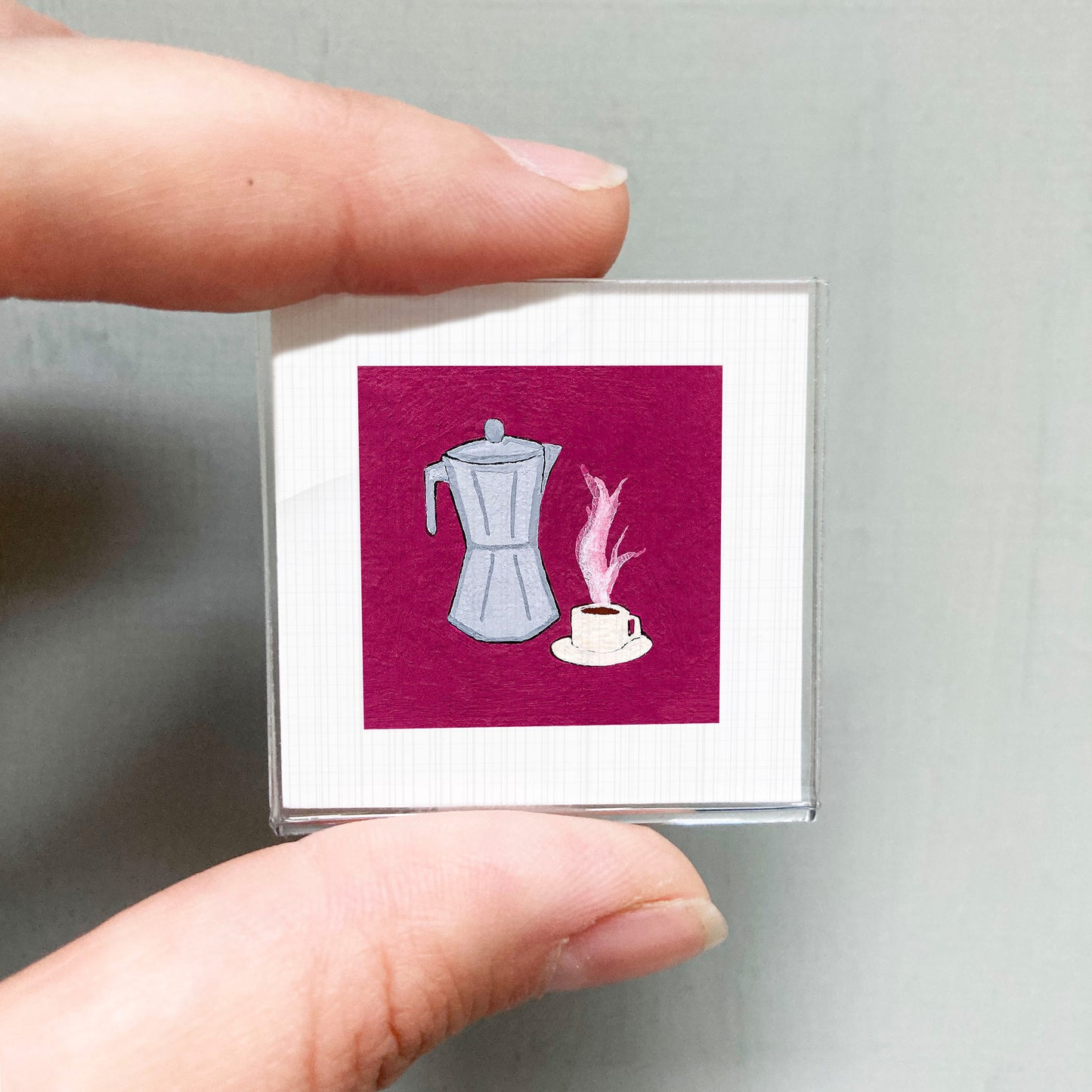 A person's hand holding a small square with an illustration of a coffee maker and a cup of coffee on it.