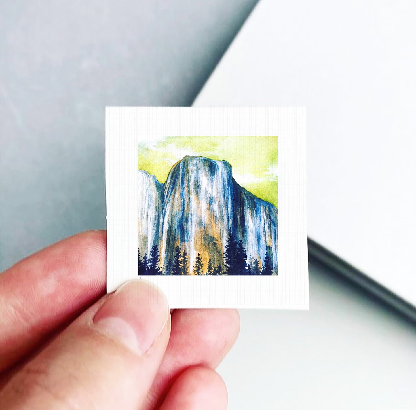 A hand is holding a small square-shaped piece of paper with a painting of a mountain landscape on it.
