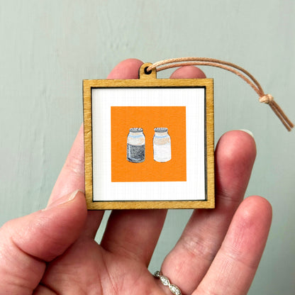 A hand holding a small wooden frame with a picture of two bottles inside it.