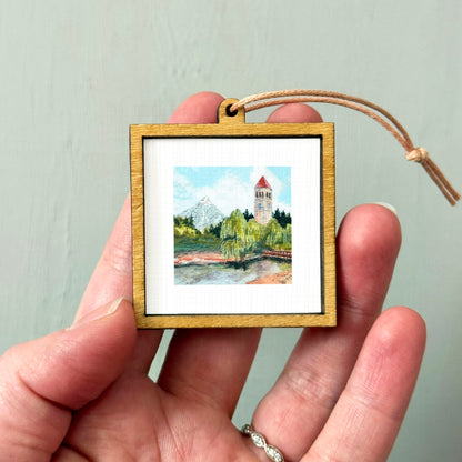 A hand holding a small wooden frame containing a painting of a mountain landscape.