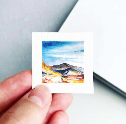 A hand is holding a small square-shaped piece of paper with a watercolor painting of a mountain landscape on it.