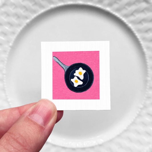 A hand is holding a small square with a pink background and a black frying pan with two white eggs on it.