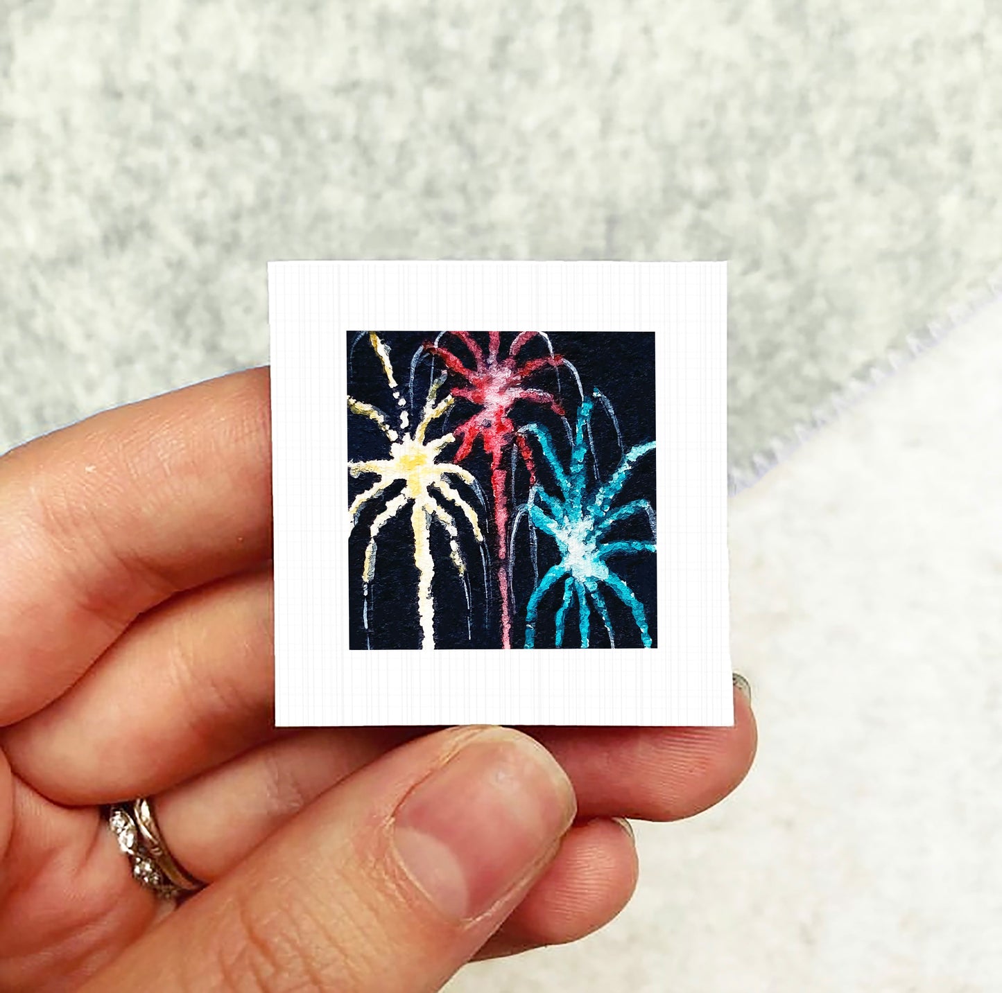 A hand holding a small square picture of colorful fireworks against a dark background.