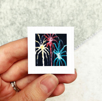 A hand holding a small square picture of colorful fireworks against a dark background.