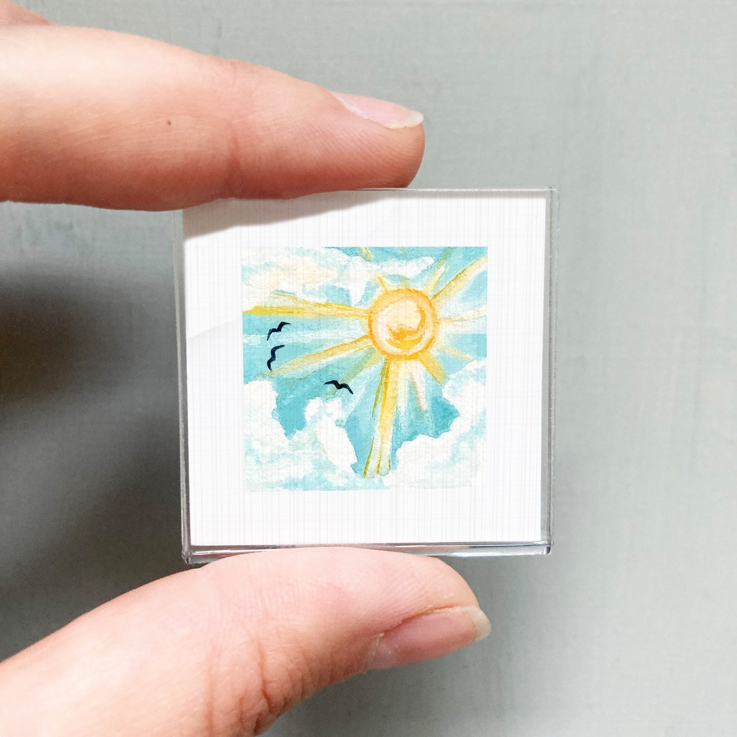 A person's hand holding a small square picture depicting a sunny sky with clouds and birds.
