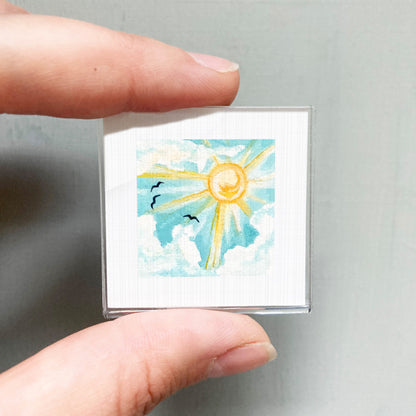 A person's hand holding a small square picture depicting a sunny sky with clouds and birds.