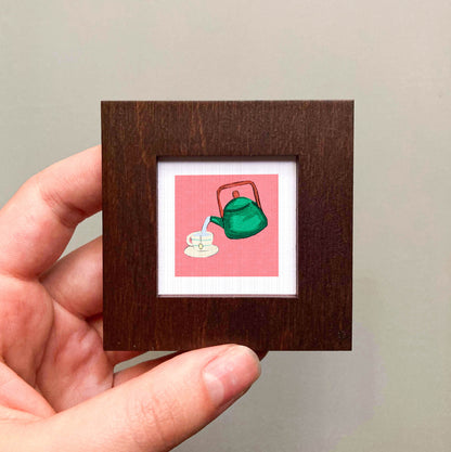 A hand is holding a small square frame containing an illustration of a green teapot pouring tea into a cup.