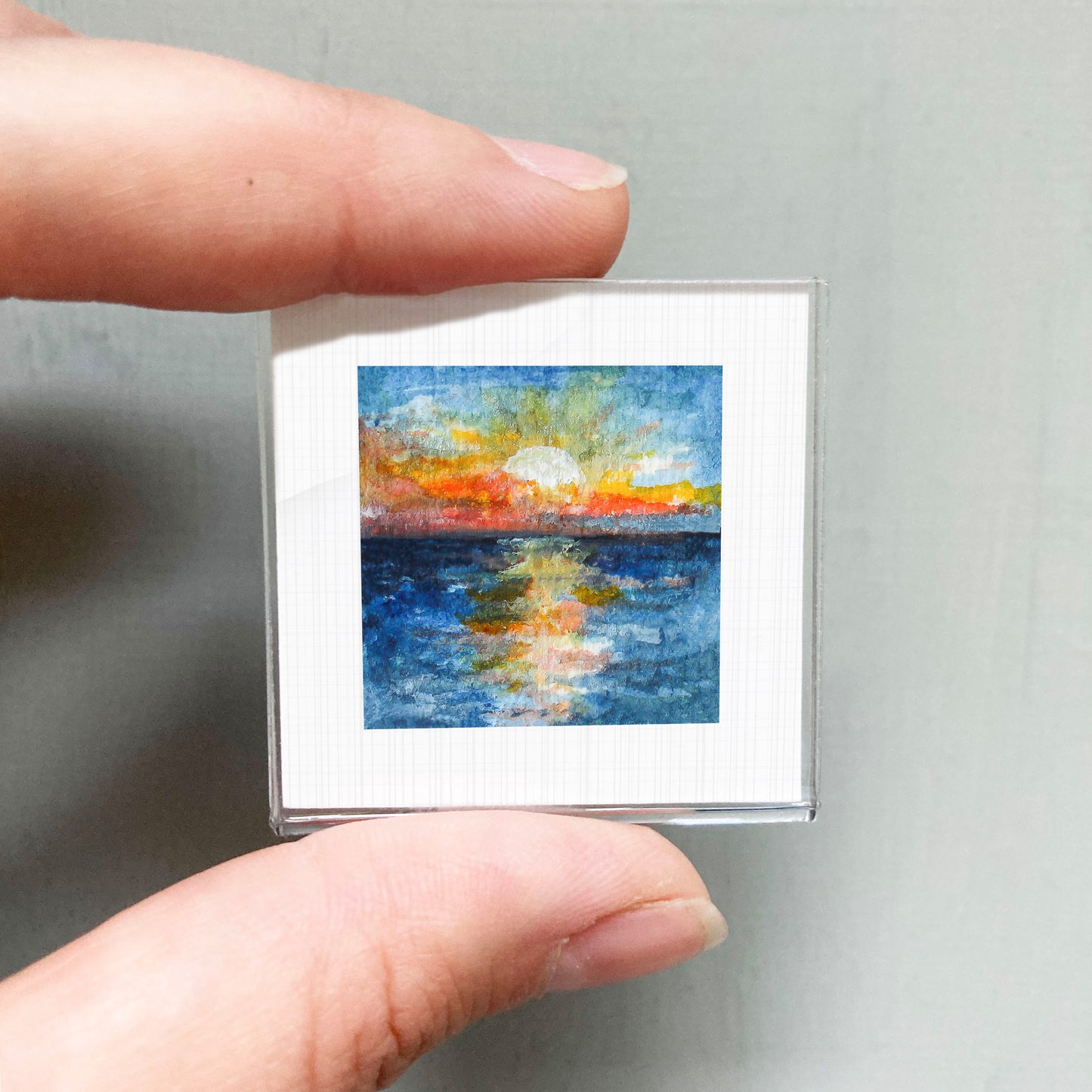 A person's hand holding a small square art print depicting a colorful sunset over a body of water.