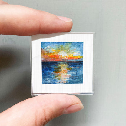 A person's hand holding a small square art print depicting a colorful sunset over a body of water.