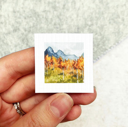 A hand holding a small square artwork depicting a mountain landscape with autumn trees.