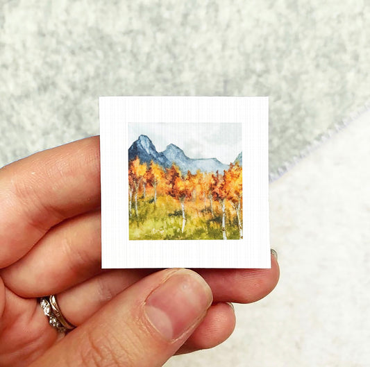 A hand holding a small square artwork depicting a mountain landscape with autumn trees.