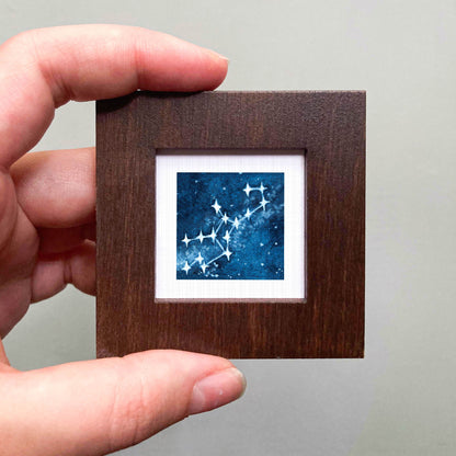 A hand is holding a small wooden frame containing a framed image of a starry night sky.