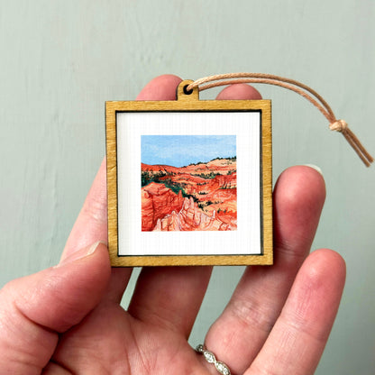A hand holding a small wooden frame containing a framed painting of a scenic landscape.