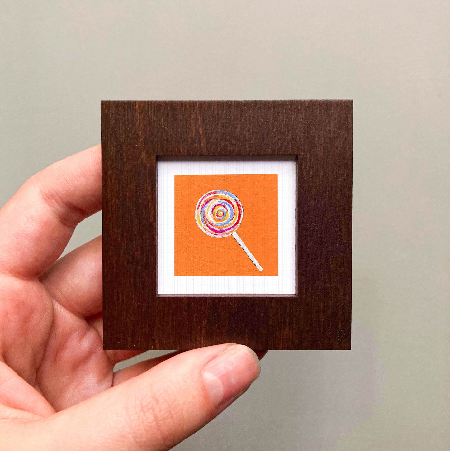 A hand is holding a small square frame containing an image of a colorful lollipop.