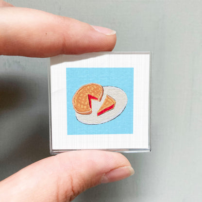 A person's hand holding a small square with an embroidered image of a piece of food, specifically a slice of pie, on a blue background.
