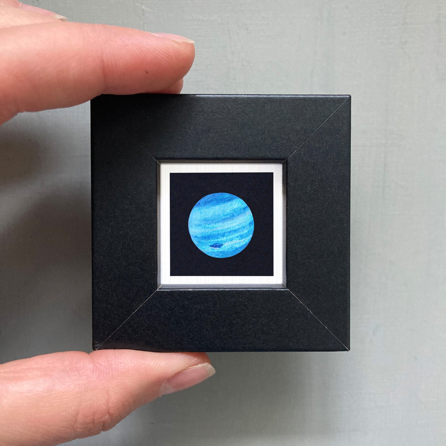 A person's hand is holding a small black square frame containing a picture of a blue planet.