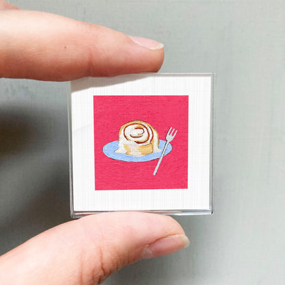 A person's hand holding a small square card with an illustration of a cinnamon roll on a blue plate, along with a fork.