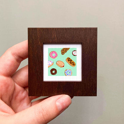 A hand is holding a small square frame containing a painting of various donuts.