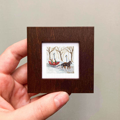 A hand is holding a small wooden frame containing a miniature painting of a horse-drawn sleigh in a snowy landscape.