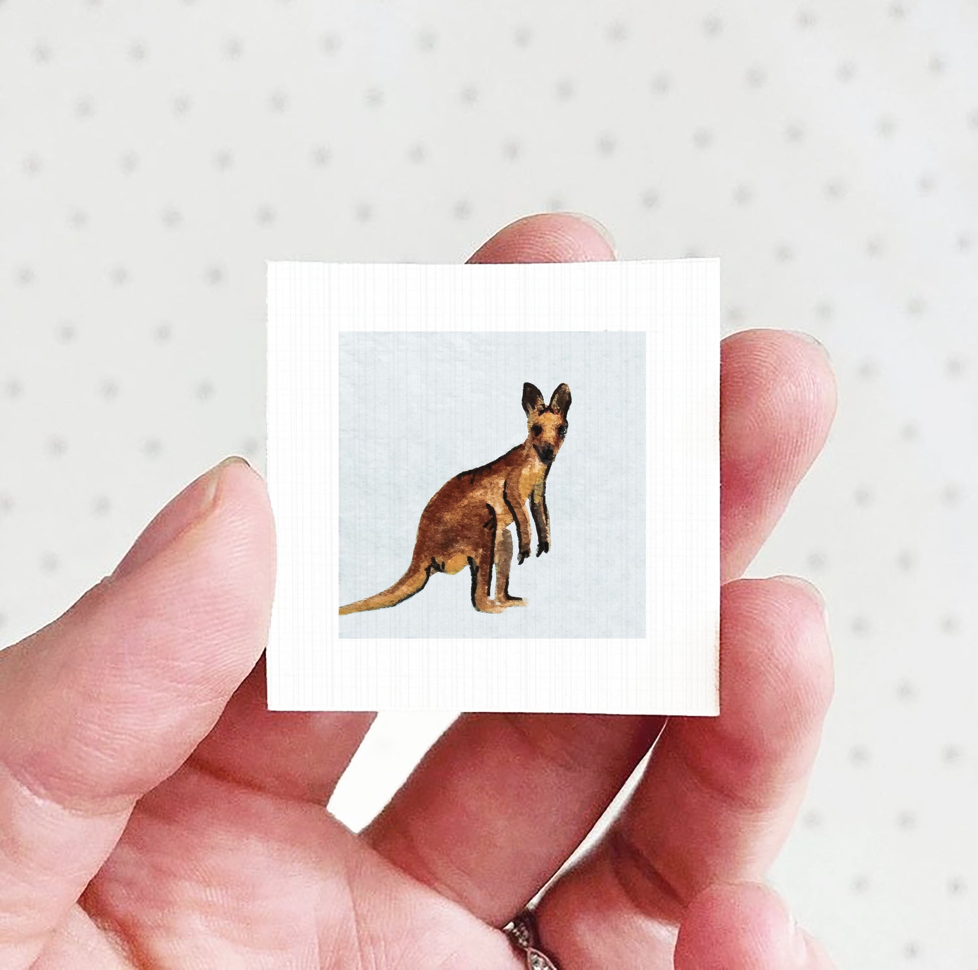 A hand holding a small square picture of a kangaroo against a white background.