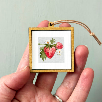 A hand holding a small wooden frame containing a framed image of a strawberry with white flowers.