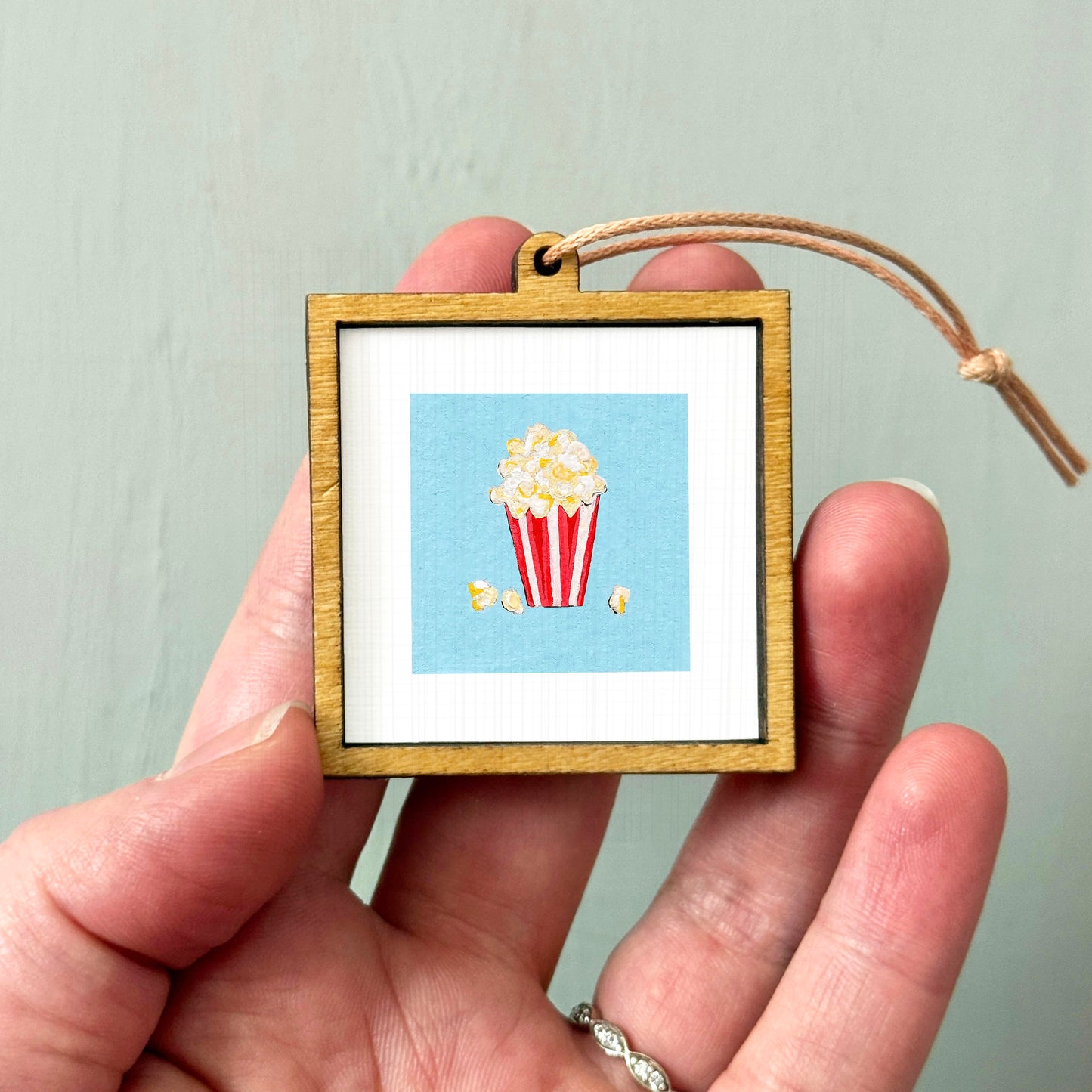 A hand holding a small wooden frame containing a picture of a red and white striped popcorn bucket.