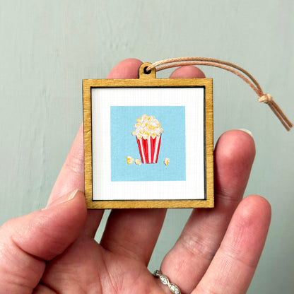 A hand holding a small wooden frame containing a picture of a red and white striped popcorn bucket.