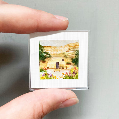 A hand holding a small square picture of a house with a garden in the background.