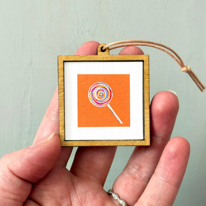 A hand holding a small wooden frame containing a colorful lollipop image.
