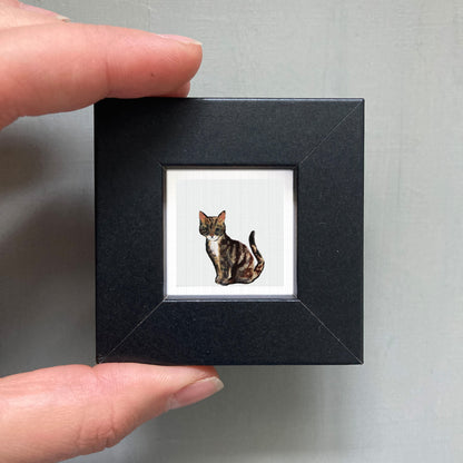 A person's hand is holding a small black picture frame containing a framed image of a cat.