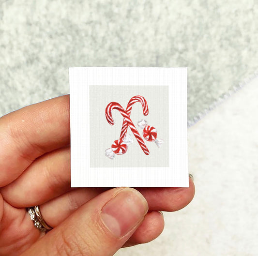 A hand holding a small square card with an illustration of two red and white striped candy canes.