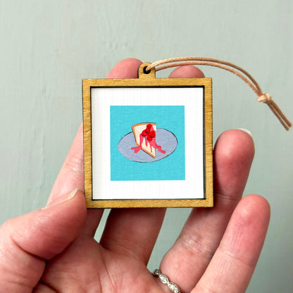 A person's hand holding a small wooden frame containing a framed illustration of a slice of pizza.