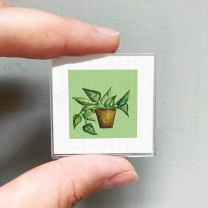 A person's hand holding a small square card with a painting of a potted plant on it.