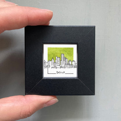 A hand is holding a small black square frame containing a watercolor illustration of the Detroit skyline.