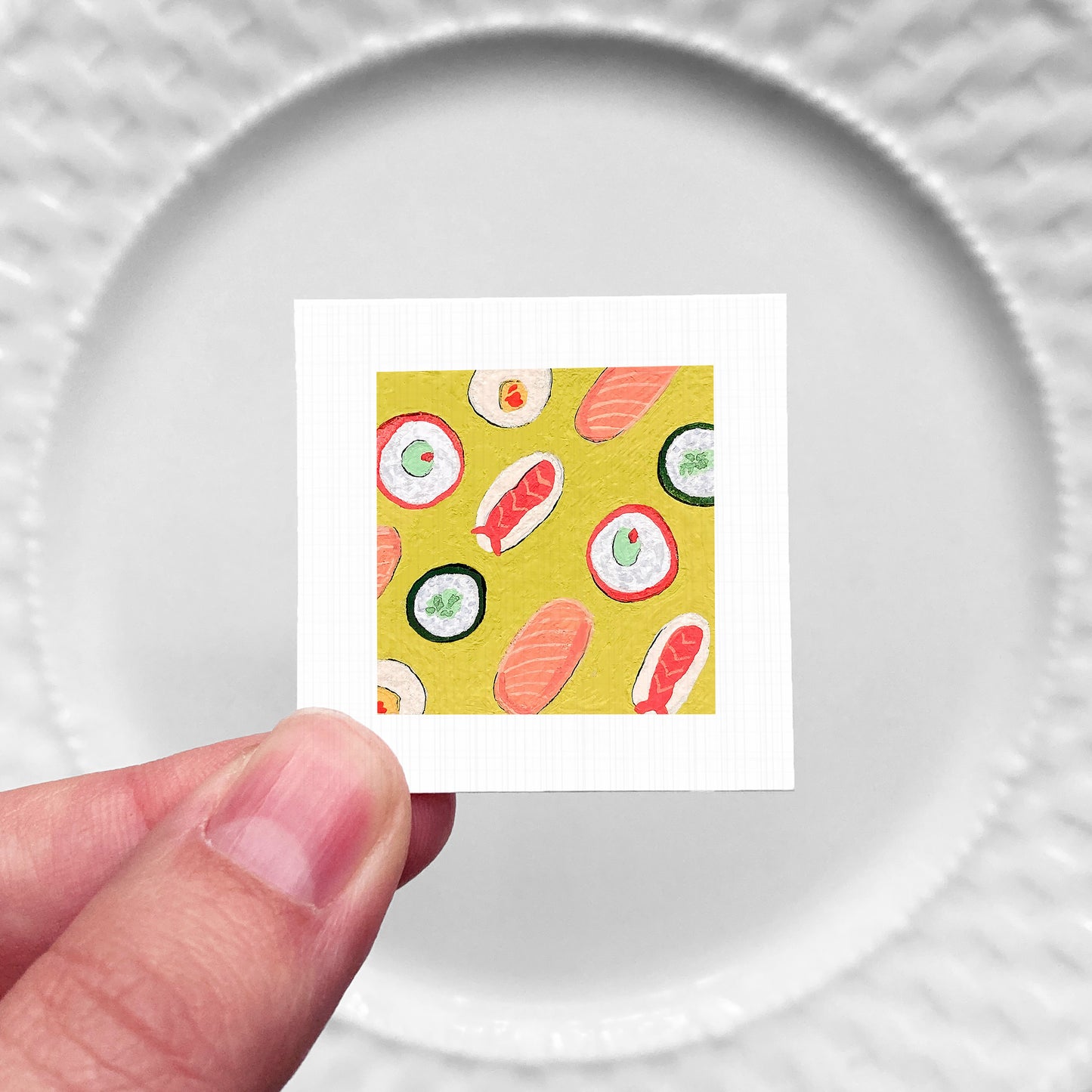 A hand is holding a small square with a vibrant illustration of sushi rolls on a yellow background.