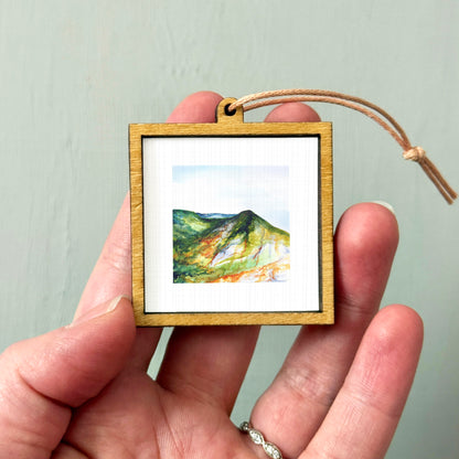 A hand holding a small wooden frame containing a watercolor painting of a mountain landscape.