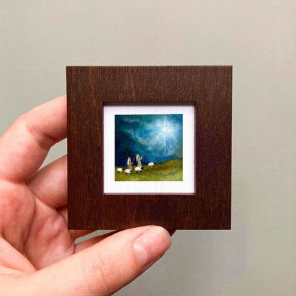A hand is holding a small, square-shaped wooden frame containing a painting of a pastoral scene with sheep grazing on a hill.