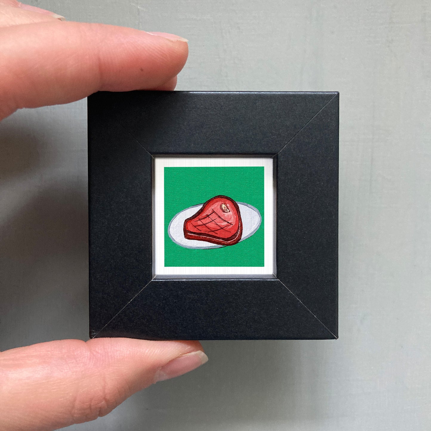 A hand is holding a small black square frame containing a vibrant red piece of meat against a green background.