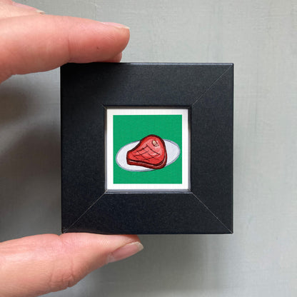 A hand is holding a small black square frame containing a vibrant red piece of meat against a green background.