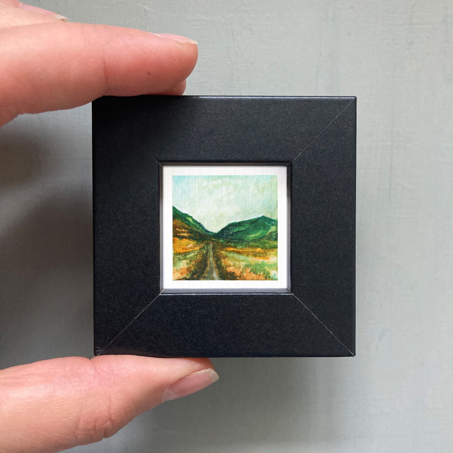 A person's hand holding a small, square-shaped black frame containing a miniature painting of a landscape with mountains and a winding road.