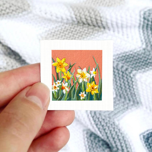 A hand is holding a small square card with a painting of yellow daffodils on it, against a background of a white and gray striped blanket.