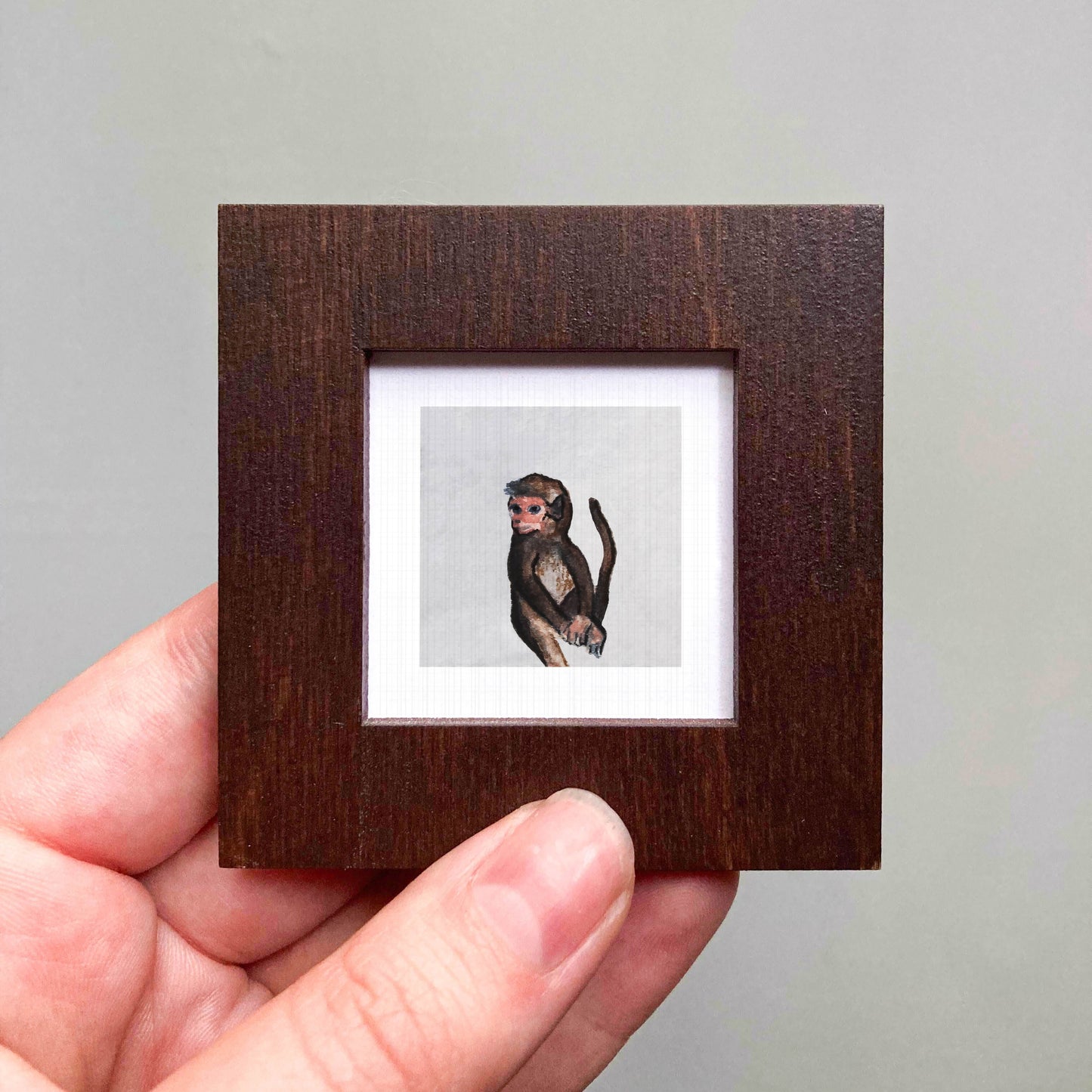 A hand is holding a small wooden frame containing a framed image of a monkey.